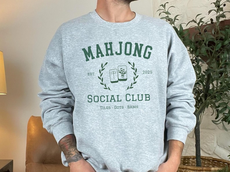 May include: A light gray sweatshirt with the words "MAHJONG SOCIAL CLUB" in green, along with the year "2025" and the words "TILES DOTS BAMS". The sweatshirt has a crew neck and long sleeves.