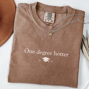 Pu&ograve; includere: Una maglietta marrone con la scritta "One degree hotter" e una grafica di cappello di laurea. Una collana d'argento con un ciondolo poggia sulla maglietta. Sono visibili anche un cappello marrone e una pianta secca.