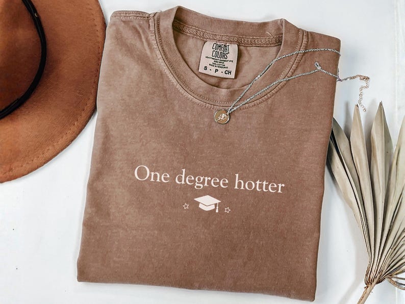 Pu&ograve; includere: Una maglietta marrone con la scritta "One degree hotter" e una grafica di cappello di laurea. Una collana d'argento con un ciondolo poggia sulla maglietta. Sono visibili anche un cappello marrone e una pianta secca.