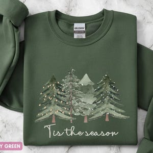 May include: A military green sweatshirt featuring a Christmas tree design and the words "Tis the season". The trees are decorated with lights, creating a festive holiday theme. The sweatshirt is a comfortable, casual garment.
