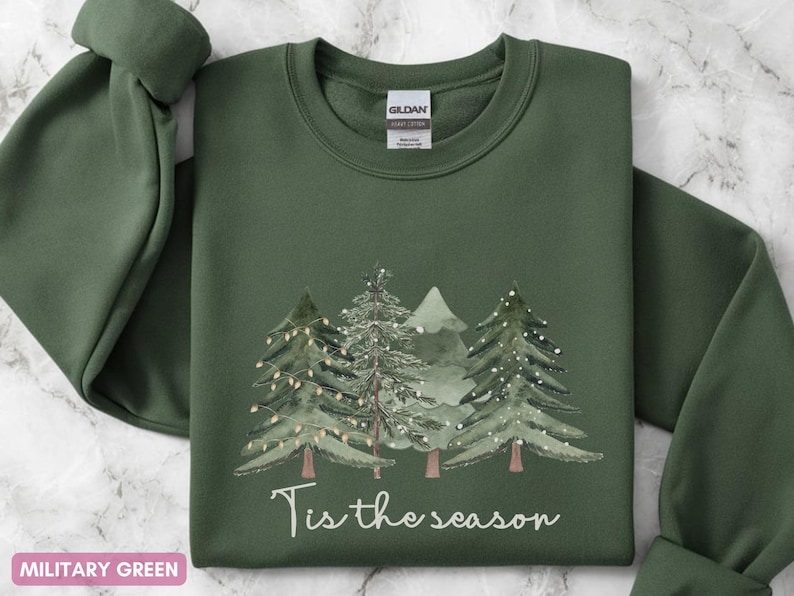 May include: A military green sweatshirt featuring a Christmas tree design and the words "Tis the season". The trees are decorated with lights, creating a festive holiday theme. The sweatshirt is a comfortable, casual garment.