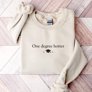 Pu&ograve; includere: Una felpa color crema con la scritta "One degree hotter" in nero, insieme a un cappello di laurea e una stella. Il top a maniche lunghe e girocollo &egrave; realizzato in un materiale morbido.