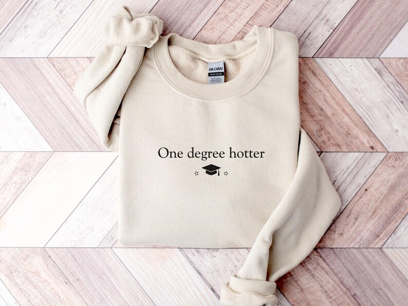 Pu&ograve; includere: Una felpa color crema con la scritta "One degree hotter" in nero, insieme a un cappello di laurea e una stella. Il top a maniche lunghe e girocollo &egrave; realizzato in un materiale morbido.