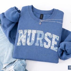 Peut inclure: Sweat-shirt bleu indigo avec le mot "NURSE" en motif floral et vichy. Un collier argenté avec un petit pendentif est posé sur le vêtement. Un jean bleu délavé est visible à gauche.