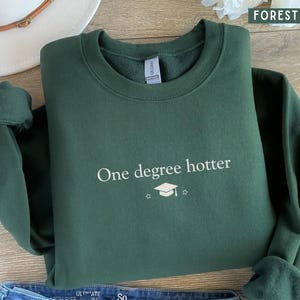 Pu&ograve; includere: Felpa verde foresta con la scritta bianca "One degree hotter" e un cappello di laurea. Sullo sfondo sono visibili un cappello bianco e jeans blu. Il colore &egrave; descritto come "Forest Green".