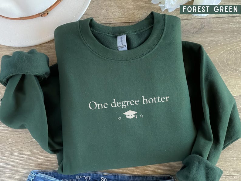 Pu&ograve; includere: Felpa verde foresta con la scritta bianca "One degree hotter" e un cappello di laurea. Sullo sfondo sono visibili un cappello bianco e jeans blu. Il colore &egrave; descritto come "Forest Green".