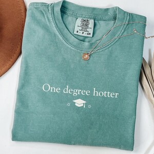 Pu&ograve; includere: Una maglietta verde acqua con la scritta "One degree hotter" e una grafica di cappello di laurea. Una collana d'oro con un ciondolo circolare poggia sulla maglietta. Un cappello marrone e una pianta secca sono sullo sfondo.