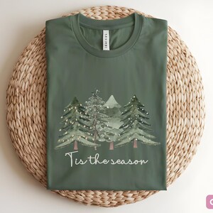 May include: Olive green t-shirt featuring a watercolour design of Christmas trees and the text "Tis the season". The shirt is folded on a woven, circular placemat.