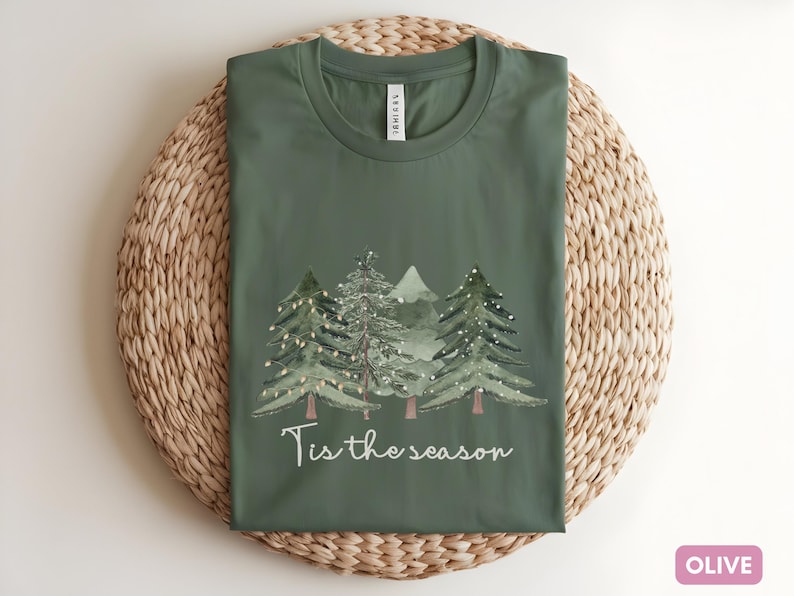 May include: Olive green t-shirt featuring a watercolour design of Christmas trees and the text "Tis the season". The shirt is folded on a woven, circular placemat.