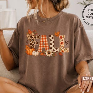 May include: An espresso-coloured short-sleeve t-shirt featuring the word FALL printed in large letters. The letters are decorated with leopard print, orange and white buffalo plaid, brown polka dots, and floral patterns, accented by pumpkins and leaves.