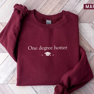 Pu&ograve; includere: Felpa bordeaux con la scritta bianca "One degree hotter", insieme a un cappello di laurea e una grafica a stella. La felpa &egrave; di colore bordeaux unito e ha uno scollo tondo.