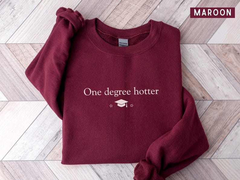 Pu&ograve; includere: Felpa bordeaux con la scritta bianca "One degree hotter", insieme a un cappello di laurea e una grafica a stella. La felpa &egrave; di colore bordeaux unito e ha uno scollo tondo.