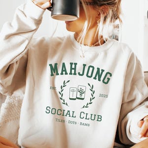 Light neutral crewneck sweatshirt featuring green collegiate-style text reading Mahjong Social Club with tile icons and decorative laurel details, styled as a cozy casual outfit for game night wear.