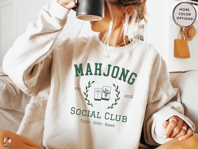 Light neutral crewneck sweatshirt featuring green collegiate-style text reading Mahjong Social Club with tile icons and decorative laurel details, styled as a cozy casual outfit for game night wear.