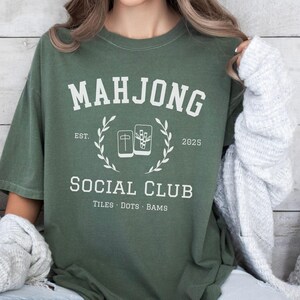 May include: Moss green t-shirt with a white graphic. The graphic reads "MAHJONG SOCIAL CLUB" with "EST. 2025" and "TILES &bull; DOTS &bull; BAMS" below. The design includes a laurel wreath and images of mahjong tiles.