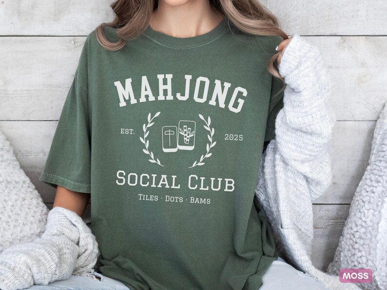 May include: Moss green t-shirt with a white graphic. The graphic reads "MAHJONG SOCIAL CLUB" with "EST. 2025" and "TILES &bull; DOTS &bull; BAMS" below. The design includes a laurel wreath and images of mahjong tiles.
