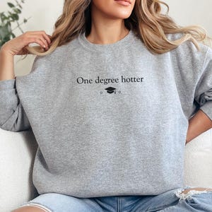 Pu&ograve; includere: Felpa grigia chiara con la scritta nera "One degree hotter", accompagnata da un cappello di laurea e una grafica a stella. La felpa &egrave; a girocollo e sembra essere realizzata in un materiale morbido.