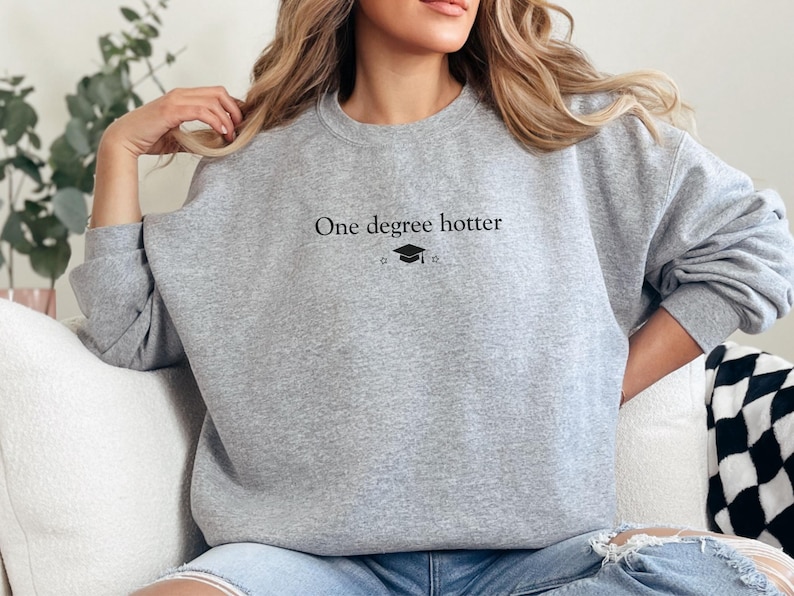 Pu&ograve; includere: Felpa grigia chiara con la scritta nera "One degree hotter", accompagnata da un cappello di laurea e una grafica a stella. La felpa &egrave; a girocollo e sembra essere realizzata in un materiale morbido.