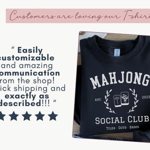 May include: Black sweatshirt with white text reading "MAHJONG SOCIAL CLUB" with a graphic of mahjong tiles. The text "EST. 2025" is also included. The sweatshirt is folded on a wooden surface.