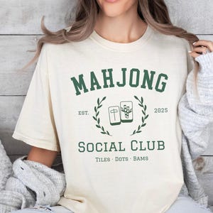 May include: Ivory-colored t-shirt with a green "MAHJONG SOCIAL CLUB" graphic. The design includes the text "EST. 2025" and illustrations of Mahjong tiles. The shirt is a casual, comfortable style.