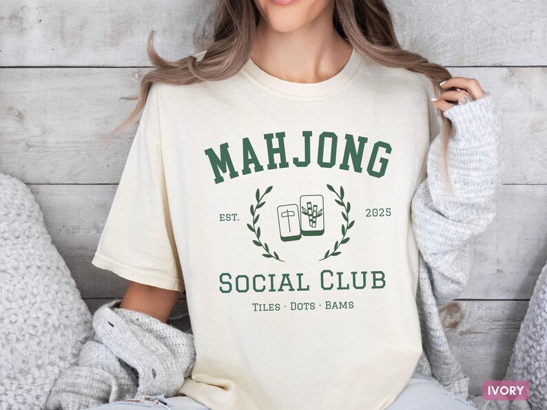 May include: Ivory-colored t-shirt with a green "MAHJONG SOCIAL CLUB" graphic. The design includes the text "EST. 2025" and illustrations of Mahjong tiles. The shirt is a casual, comfortable style.