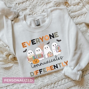 May include: White sweatshirt with the text "EVERYONE COMMUNICATES DIFFERENTLY" and illustrations of ghosts, bats, a spiderweb, and a pumpkin. The sweatshirt has long sleeves and a crew neck.