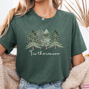 May include: Dark green t-shirt featuring a watercolour illustration of three Christmas trees and the words "Tis the season". The trees are adorned with small white lights. A festive design for the holiday season.