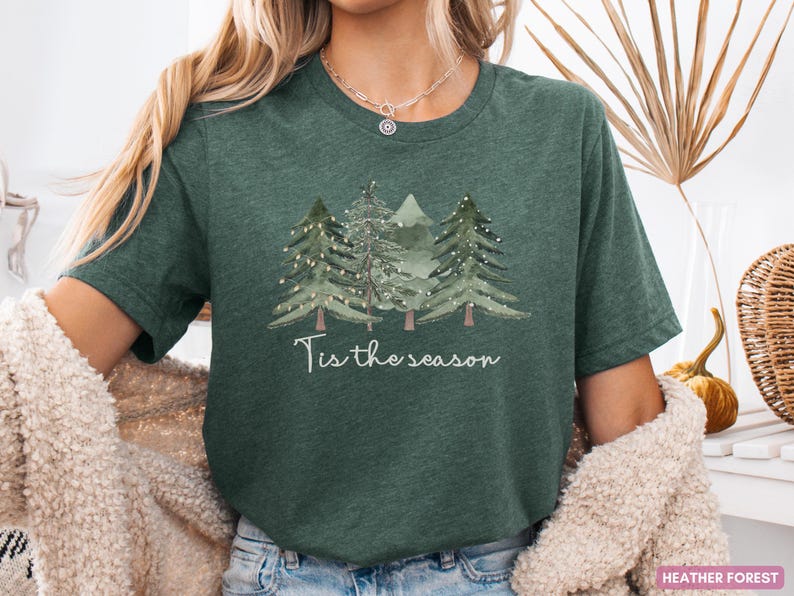 May include: Dark green t-shirt featuring a watercolour illustration of three Christmas trees and the words "Tis the season". The trees are adorned with small white lights. A festive design for the holiday season.
