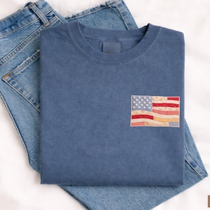 May include: A blue t-shirt with a small American flag patch on the chest, folded on top of a pair of blue jeans. The flag patch features red and white stripes and a field of white stars on a blue background. The word "DENIM" is visible.