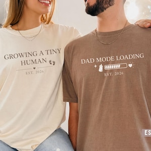 May include: Two matching t-shirts, one cream and one espresso. The cream shirt says "Growing a Tiny Human" with a footprint graphic and "Est. 2026." The espresso shirt says "Dad Mode Loading" with a loading bar and "Est. 2026."