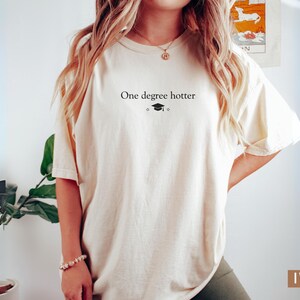 Pu&ograve; includere: T-shirt color avorio con la scritta "One degree hotter" e un cappello di laurea. La maglietta ha una vestibilit&agrave; ampia e maniche corte. La parola "IVORY" &egrave; stampata in un rettangolo marrone in basso a destra.