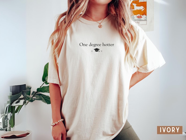 Pu&ograve; includere: T-shirt color avorio con la scritta "One degree hotter" e un cappello di laurea. La maglietta ha una vestibilit&agrave; ampia e maniche corte. La parola "IVORY" &egrave; stampata in un rettangolo marrone in basso a destra.