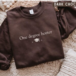 Pu&ograve; includere: Felpa marrone cioccolato scuro con la scritta bianca "One degree hotter", insieme a un cappello di laurea e una grafica a stella. La felpa &egrave; esposta su una superficie strutturata.