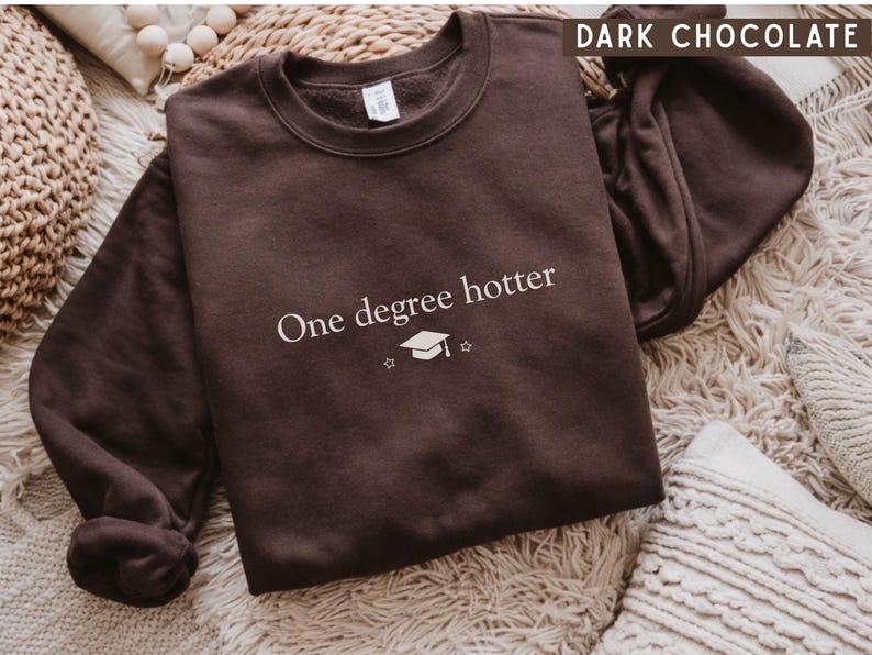 Pu&ograve; includere: Felpa marrone cioccolato scuro con la scritta bianca "One degree hotter", insieme a un cappello di laurea e una grafica a stella. La felpa &egrave; esposta su una superficie strutturata.