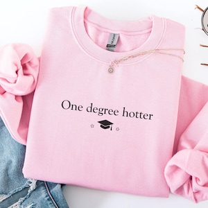 Pink sweatshirt with minimalist “one degree hotter” text and graduation cap graphic, designed for masters graduates, cozy long sleeve pullover for college graduation celebration and everyday wear.