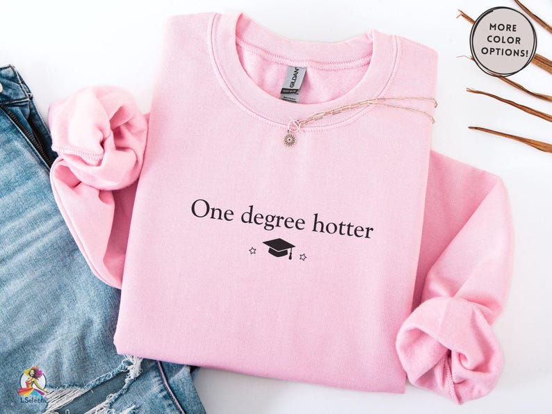 Pink sweatshirt with minimalist “one degree hotter” text and graduation cap graphic, designed for masters graduates, cozy long sleeve pullover for college graduation celebration and everyday wear.