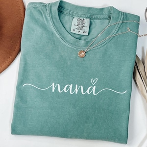 May include: A seafoam green t-shirt with the word "nana" in a cursive font and a small heart above the "a". A gold necklace with a pendant rests on the shirt. The shirt has a Comfort Colors tag.