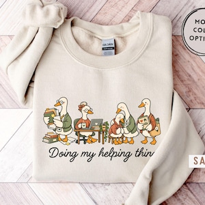 May include: A sand-coloured crewneck sweatshirt featuring a graphic of four cartoon ducks engaged in various activities, including reading, working on a laptop, and holding a book. The text below the illustration reads, Doing my helping thing.