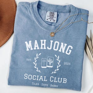 May include: A light blue Comfort Colors t-shirt with the words "MAHJONG SOCIAL CLUB" in white. The shirt features a graphic of Mahjong tiles and the text "TILES DOTS BAMS". A gold necklace and a brown hat are also in the image.