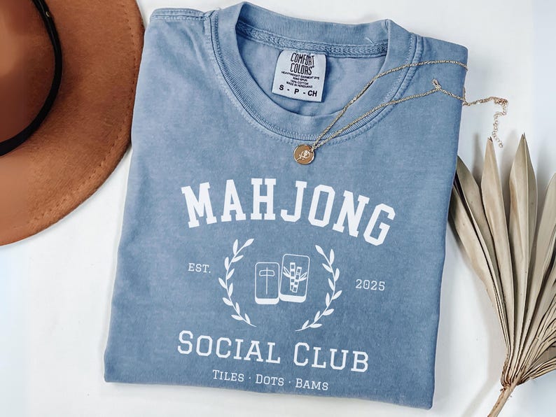 May include: A light blue Comfort Colors t-shirt with the words "MAHJONG SOCIAL CLUB" in white. The shirt features a graphic of Mahjong tiles and the text "TILES DOTS BAMS". A gold necklace and a brown hat are also in the image.
