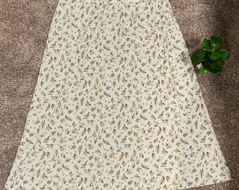 May include: A cream-colored midi skirt with a floral pattern in shades of brown and green. The skirt has a ribbed texture and a slight A-line shape. The skirt is a versatile piece for a casual or semi-formal outfit.