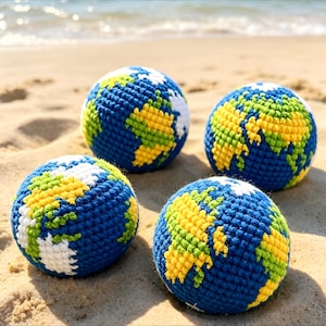 Puede incluir: Cuatro bolas de ganchillo con forma de globo terráqueo sobre una playa de arena. Las bolas presentan océanos azules y continentes blancos, amarillos y verdes. Las bolas están dispuestas en grupo.