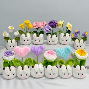 May include: A collection of handmade crochet flower arrangements. Each arrangement features a white bunny-shaped pot with a variety of colorful crochet flowers and heart-shaped designs. The flowers include yellow, pink, purple, and blue blooms.