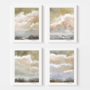 May include: Four framed watercolor prints featuring cloudscape art in muted tones of beige, pink, and gray. Each print has a white border and a signature in the bottom right corner. The artwork depicts a landscape with a river.