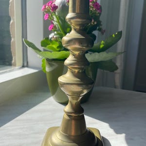 Puede incluir: Un candelero de latón con base cuadrada y diseño decorativo escalonado. El candelero está frente a una ventana con una planta en maceta con flores rosas.