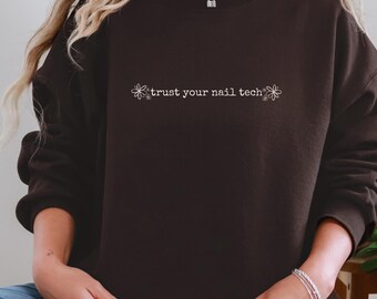 Op de afbeelding: Een donkerbruine sweatshirt met de witte tekst "trust your nail tech", geflankeerd door bloemmotieven. De sweatshirt is effen van kleur en heeft een ronde hals.