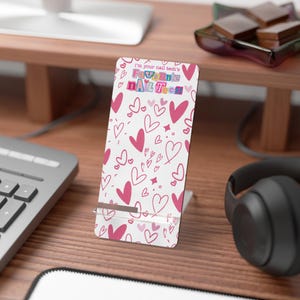 Könnte beinhalten: Ein weißer Telefonständer mit einem rosa Herzmuster und dem Text "I'm your nail tech's Favorite NAIL Tech". Der Ständer steht auf einem Holzschreibtisch mit Tastatur und Kopfhörern im Hintergrund.
