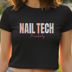 May include: Black t-shirt with the words "NAIL TECH" in a floral pattern and the name "Kimberly" in pink script. The shirt is a crew neck and appears to be made of a soft material.