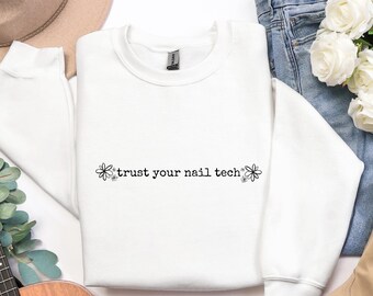 Op de afbeelding: Een witte crewneck sweatshirt met de zwarte tekst "trust your nail tech", versierd met bloemmotieven. De sweatshirt wordt gepresenteerd tegen een witte achtergrond, met een blauwe jeans en een boeket witte rozen op de achtergrond.