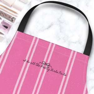 May include: Pink apron with vertical white stripes and a black neck strap. The apron has the text "I do all the Pretty Girls Nails" in a cursive font. Nail care tools are visible in the background.